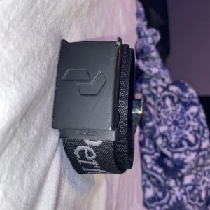 Svart textilbälte från Peak  - Säljer ett svart bälte från Peak med metallspänne och präglad logga på spännet. Bältet är gjort i slitstark textil och har vit text längs bandet. Perfekt accessoar för att lyfta din outfit.