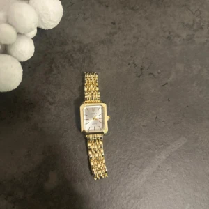 Guldfärgad klocka med länkarmband - Snygg guldfärgad klocka  med rektangulärt ur och länkarmband i metall. Urtavlan är vit med guldiga visare och markeringar, vilket ger en clean och modern vibe. Perfekt accessoar för dig som gillar stilrena detaljer och vill sticka ut.