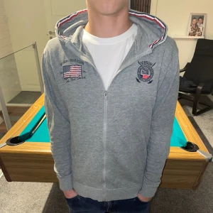 Grå hoodie från Vinson Polo Club - Snygg grå hoodie från Vinson Polo Club med dragkedja och broderade loggor på bröstet. Huva med röd, vit och blå randig insida ger en sportig touch. Mjuk bomullsblandning och två fickor framtill. Perfekt för chill dagar.