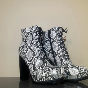 Svarta och vita boots med ormmönster - Säljer ett par boots med chunky hög svart klack och snörning, täckt i svartvitt ormmönster. Skorna har grov sula för extra grepp och dragkedja på insidan för enkel påtagning. Perfekt statement piece för dig som vill sticka ut.