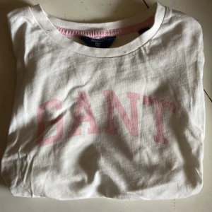 Vit GANT t-shirt med rosa tryck - Vit t-shirt från GANT med klassiskt rosa logotyp-tryck framtill. Rund hals med rosa ribbad detalj på insidan av kragen. Tillverkad i mjuk bomull, perfekt för en avslappnad och stilren look.