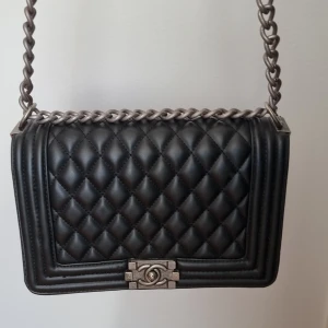 Svart quiltad Bag - Säljer en ikonisk svart Bag i quiltat skinn med silverfärgad kedjerem och klassiskt CC-lås framtill. Väskan har en boxig form och coola detaljer i metall. Perfekt för dig som vill ha en statementväska med lyxig känsla och tidlös design.