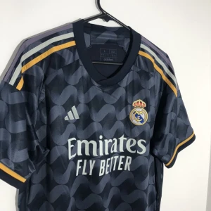 Real Madrid bortatröja Adidas  - Real Madrid bortatröja 23/24  från Adidas i mörkblått med vågigt mönster, gula detaljer och klubbmärke på bröstet. Produkten är i storlek S men kommer passa dig som är 175-180cm. T-Shirten är i nytt skick och helt oanvänd.       Nypris: 1200kr