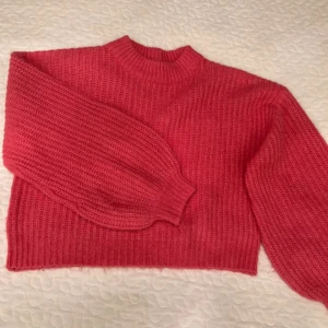 Rosa stickad tröja från Vila - Superfin rosa stickad tröja från Vila med ribbad struktur och hög hals. Tröjan har långa ärmar med breda muddar och en lite boxig, croppad passform. Perfekt för att addera färg till din outfit!