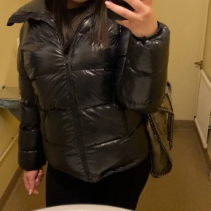 Glansig pufferjacka svart - Svart glansig pufferjacka i strl 40 (L) men sitter som M. Köpt förra vintern och säljer då den ej passar längre, jackan har ett hål vid handleden men annars är den i bra skick. Den är väldigt varm med stor luva, perfekt för kalla vinter dagar (från någon som fryser mycket). Från Everest, nypris 1299kr. 