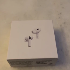 AirPods Pro (2nd generation) - Apple AirPods Pro (2nd generation) i originalförpackning med laddningsetui och kabel. Produkten ser ut att vara i mycket gott skick, komplett med tillbehör och kartong. Perfekt för brusreducering och trådlös musikupplevelse.