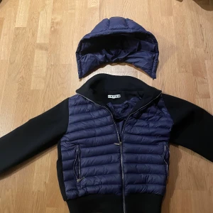 Cardigan - Säljer en snygg cardigan från Levon i mörkblått och svart. Jackan har avtagbar huva, dragkedja framtill och två fickor med dragkedja. Ärmarna är svarta och kroppen är quiltad i blått. Perfekt för vinter och höst, inte många såna i storlek xs, Ny pris 1500, och finns inte längre att köpa. Hör gärna av er vid funderingar!