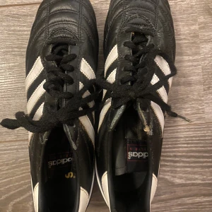 Adidas Kaiser 5 fotbollsskor svart/vit - Klassiska Adidas Kaiser 5 fotbollsskor i svart skinn med vita ränder och snörning. Skorna har rund tå och metall-dobbar för bästa grepp på planen. Perfekta för dig som vill ha en tidlös och stilren look på fotbollsplanen.