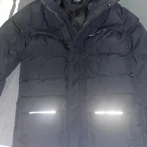 Svart pufferjacka från Everest 164 - Svart pufferjacka från Everest med reflekterande detaljer vid fickorna och bröstet. Jackan har en broderad Everest-patch på ärmen och dragkedja framtill. Perfekt för kalla dagar med sin vadderade design och höga krage.