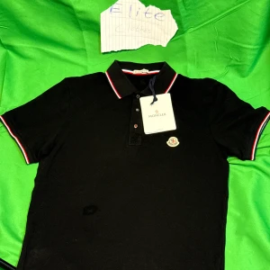 Svart Moncler pikétröja med röd/vit kant - Svart pikétröja  Moncler med klassisk krage och knappar framtill. Röd och vit rand längs krage och ärmslut ger en snygg kontrast. Moncler-logga på bröstet. Perfekt för dig som vill ha en stilren och exklusiv look.Liknande modell som originalet!