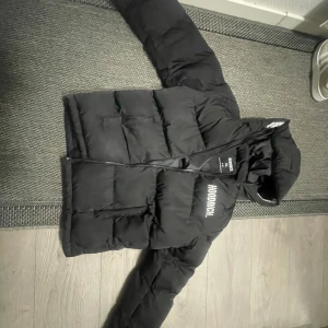 Svart pufferjacka från Hoodrich XL - Svart pufferjacka från Hoodrich i storlek XL, med tydlig logga på bröstet och huvan. Jackan har dragkedja framtill, två fickor och en vadderad look som ger en riktigt street och modern vibe. Perfekt för kalla dagar och snygg till sneakers.