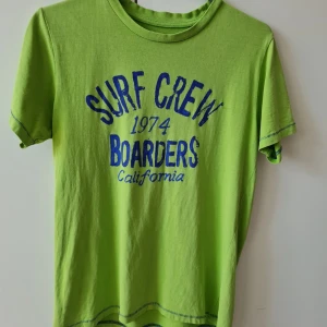 T-shirt från Cubus strl 164 (barn) - Säljer en grön t-shirt med blå tryck 'Surf Crew 1974 Boarders California' framtill. T-shirten har korta ärmar och rund hals. Perfekt för en avslappnad stil och somrig känsla. 100% bomull. Storlek 164. Kan hämtas i Vallentuna.