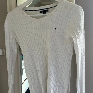 Vit kabelstickad tröja Tommy Hilfiger - Vit kabelstickad tröja från Tommy Hilfiger i klassisk modell med rund halsringning och diskret logga på bröstet. Långärmad och i mjuk bomull, perfekt för lager-på-lager och snygg till jeans eller kjol. Tröjan har en liten gulaktig fläck (se bild 3) syns knappt men därav priset