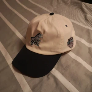 keps Authentic Brand - Snygg keps i beige med mörk skärm från Authentic Brand. Framtill finns ett broderat A och på sidan en basketinspirerad patch med texten 'Authentic Brand'. Justerbar rem baktill i metall. Perfekt accessoar för en sportig look.