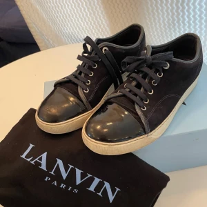 Svarta sneakers från Lanvin Paris - Lanvin sneakers i svart mocka med glansig svart tå i skinn och vita gummisulor. Klassisk rund tå, snörning och diskreta sömmar. Snygg kontrast mellan material och färg, perfekt för dig som gillar stilrena detaljer. De kommer med orginalboxen och original dustbag och kostar 1599kr. Skorna är i storlek 41/42
