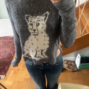 Grå stickad tröja med leopardmotiv Hollister - Mysig grå stickad tröja från Hollister med ett stort, ljust leopardmotiv framtill. Tröjan har lång ärm, rund halsringning och en något croppad passform. Perfekt för dig som vill ha en cool och bekväm look med lite edge.