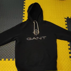 Blå hoodie från GANT med logga - Blå Mork hoodie från GANT med vit broderad logga på bröstet och vita snören i huvan. Klassisk modell med känguruficka och mjukt material, perfekt för chill dagar. Snygg och enkel att matcha med jeans eller joggers.