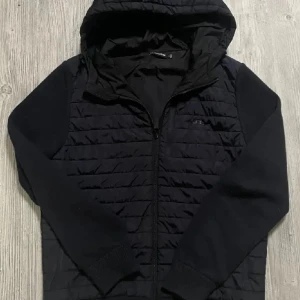 Svart quiltad jacka från J.Lindeberg - Svart quiltad jacka från J.Lindeberg med huva och dragkedja. Jackan har stickade ärmar och vadderad kropp, vilket ger en snygg kontrast. Perfekt för kyliga dagar och har en stilren look med diskret logga på bröstet.