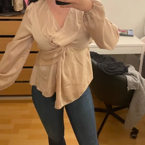 BLUS i satin - Säljer en champagnefärgad omlottblus med lång ärm och knyt framtill. Blusen har v-ringning, lätt glansigt satintyg och en lös passform som ger en snygg siluett. Perfekt att styla med jeans eller kjol för en trendig look. Aldrig använd.  Satin