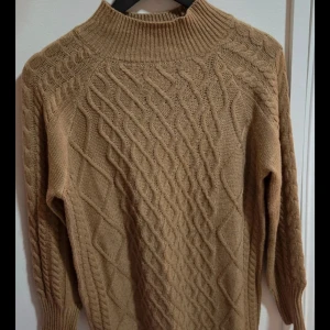 Stickad Tröjklänning Polokrage | Kabelstickad Oversized Kamelfärg Beige - Säljer en jättemysig och varm tröjklänning i en fin neutral beige/kamelfärgad nyans. Klänningen har en hög polokrage och ett vackert, varierat flätstickat mönster (cable knit). Perfekt för höst och vinter, och kan bäras med strumpbyxor och stövlar eller över leggings. Stilen är lite oversized/lös i passformen, vilket gör den extra bekväm.  Färg: Neutral Beige / Ljusbrun (Kamelfärg) Detaljer: Polokrage, Flätstickat/kabelstickat mönster.  Stil: Oversized/lös passform. Mått: Längd: 83 cm, Axel: 41 