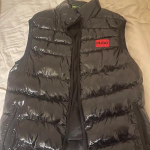 Svart dunväst från Hugo - Snygg svart dunväst från Hugo med glansig finish och stora röda logotyper både fram och bak. Västen har dragkedja och två fickor med dragkedja på sidorna. Perfekt för lager-på-lager och ger en cool streetstyle-vibe.