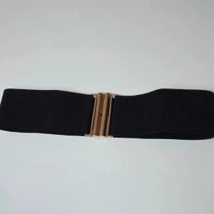 Svart brett bälte med guldfärgat spänne - Snyggt svart bälte med bredd och elastiskt material, perfekt för att ge extra edge till din outfit. Bältet har ett stilrent guldfärgat metallspänne med geometrisk design som ger en modern touch. Passar till klänning, kjol eller över en skjorta för att markera midjan.