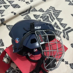Blå hockeyhjälm CCM med galler - Säljer en mörkblå hockeyhjälm från CCM med silverfärgat galler och svart hakskydd. Hjälmen har justerbara remmar och vadderad insida för extra komfort. Perfekt för dig som spelar hockey och vill ha en stilren och säker hjälm.