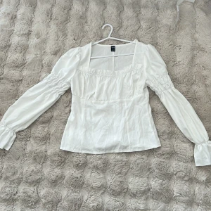 Vit blus med puffärmar från SHEIN - Supersöt vit blus från SHEIN i storlek S med puffiga långärmar och smockdetaljer vid ärmarna. Blusen har fyrkantig ringning och är figursydd i midjan. Perfekt för dig som gillar romantisk och trendig stil.