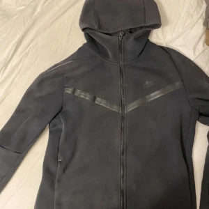 Svart Nike hoodiejacka med dragkedja M - Svart hoodiejacka från Nike i storlek M. Jackan har hel dragkedja, huva med snörning och diskret Nike-logga på bröstet. Snygg V-detalj i svart på framsidan. Mjuk och bekväm, perfekt för chill eller träning. Liten defekt på sista bilden. 
