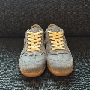 Maison Margiela sneakers - Maison Margiela sneakers i grå mocka och skinn med beige snörning och gummisula. Klassisk siluett med rund tå och platt sula. Snygga detaljer och stilren design som passar till allt. Märke och storlek syns på sulan.