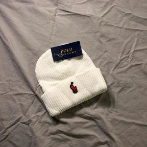 Vit ribbad mössa från Polo Ralph Lauren - Vit ribbstickad mössa från Polo Ralph Lauren med klassisk uppvikt kant och röd broderad logga framtill. Perfekt accessoar för kyliga dagar och enkel att matcha med olika outfits. Skickas samma dag och pris kan diskuteras vid snabb affär!