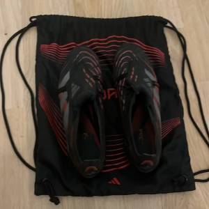 Adidas Predator fotbollsskor svart/röd - Säljer ett par Adidas Predator fotbollsskor i svart med röda och grå detaljer. Skorna har snörning, låg profil och mönstrad ovandel för extra grepp på planen. Snygg design med klassiska tre ränder på sidan och texten Predator. Perfekta för dig som vill sticka ut på fotbollsplanen.