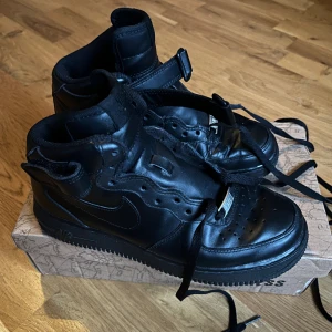 Nike Air Force 1 High Svarta Skinn - Säljer ett par svarta Nike Air Force 1 High med klassisk siluett, snörning och kardborreband runt ankeln. Skorna är helt i svart skinn med perforerad tå och Nike-logga på sidan. Perfekta för dig som gillar streetstyle och vill ha en ikonisk sneaker i garderoben.