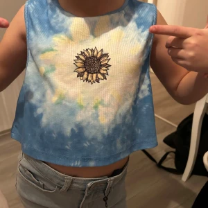 Blå tie-dye croppad linnetopp med solros - Säljer en croppad ärmlös linnetopp i blå och vit tie-dye med broderad solros framtill. Toppen har rund halsringning och är perfekt för varma dagar. Skön och lätt att bära, passar till jeans eller shorts.