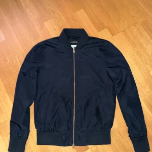 J.Lindeberg marinblå bomberjacks (S) - Snygg mörkblå bomberjacka från J.Lindeberg med klassisk passform. Jackan har dragkedja framtill, ribbade muddar och krage samt diskret logga på insidan. Perfekt för dig som gillar stilren och enkel design.