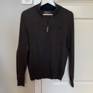 Mörkbrun merinoullströja Morris S - Snygg mörkbrun stickad tröja från Morris i 100% merinoull. Tröjan har half zip-dragkedja med en läderdetalj, krage och lång ärm. Perfekt för dig som gillar stilrena och klassiska plagg med en modern touch. En liten defekt finns på lappen i nacken där en tråd har lossnat i ett hört, men detta kan sys fast väldigt enkelt. 
