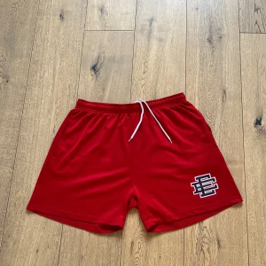Röda shorts från Eric Emanuel XL - Röda shorts från Eric Emanuel i storlek XL. De har vit snörning i midjan och ett stort EE-logotyptryck på ena benet. Shortsen är tillverkade i ett lätt och sportigt syntetmaterial, perfekta för sommaren eller träning.