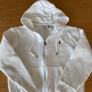 Vit hoodie från Polo Ralph Lauren - Vit hoodie från Polo Ralph Lauren med dragkedja och huva. Klassisk design med broderad logga på bröstet, långa ärmar och fickor framtill. Tillverkad i mjuk bomull, perfekt för en clean och stilren look.