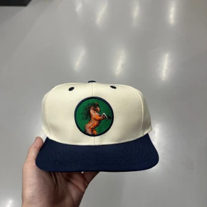 Beige keps med hästbrodyr - Snygg beige keps med mörkblå skärm och broderad häst på grön bakgrund framtill. Justerbar snapback baktill och grön undersida på skärmen. Perfekt accessoar för dig som gillar unika detaljer och sportig stil.