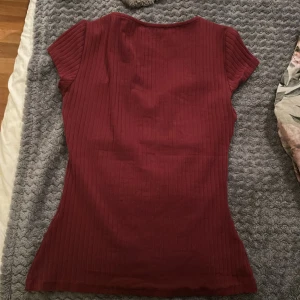 Vinröd ribbad topp från Anna Field - Säljer en vinröd ribbad topp från Anna Field med korta ärmar och snygg v-ringning. Toppen har en figurnära passform och är tillverkad i mjuk bomullsmix. Perfekt för dig som gillar stilrena och bekväma plagg med lite edge.