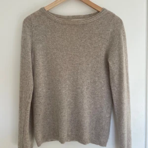 Beige stickad tröja i ull - Mysig beige stickad tröja med rund hals och långa ärmar. Tröjan är i mjuk ull och har en enkel, klassisk design med ribbade muddar vid ärmslut och nederkant. Perfekt för lager-på-lager under kyliga dagar.