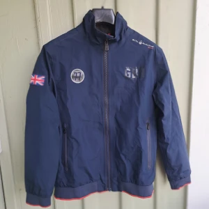 Blå Sail Racing Gore-Tex vindjacka - Snygg marinblå vindjacka från Sail Racing med Gore-Tex-material. Jackan har hel dragkedja, hög krage, Union Jack-flagga på ärmen och coola tryckdetaljer. Perfekt för dig som gillar sportig stil och vill hålla dig torr och varm även när det blåser och regnar.