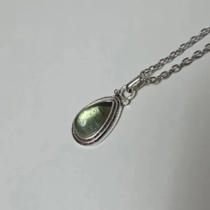 Silverfärgat halsband med grön droppe - Stilrent halsband i silverfärg med en kedja och ett droppformat hänge. Hänget har en glansig, ljusgrön sten som skimrar snyggt i ljuset. Perfekt för dig som gillar enkla men unika smycken med en touch av färg.