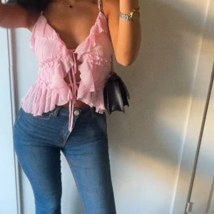 Rosa volangig linnetopp med knyt - Supersöt rosa linnetopp med tunna axelband, volangdetaljer och knyt framtill. Toppen har ett lätt plisserat tyg och är croppad med en luftig känsla. Perfekt för varma dagar och ger en romantisk vibe till din outfit.