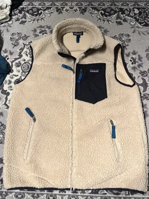 Beige fleeceväst från Patagonia - Säljer en beige fleeceväst från Patagonia med marinblå detaljer och dragkedjor. Västen har en hög krage, tre fickor med blå dragkedjor och en snygg bröstficka i marinblått tyg. Perfekt för lager-på-lager under kyliga dagar.