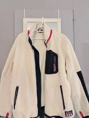 Vit teddyjacka från GANT XL - Säljer en vit teddyjacka från GANT i storlek XL. Jackan har svarta detaljer, dragkedjor och en bröstficka med GANT-logga. Röd rand på kragen och patch nertill. 