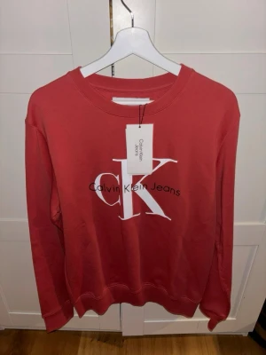 Röd sweatshirt från Calvin Klein Jeans - Säljer en röd sweatshirt från Calvin Klein Jeans med stort vitt CK-tryck. Aldrig använd, prislappen är kvar! 