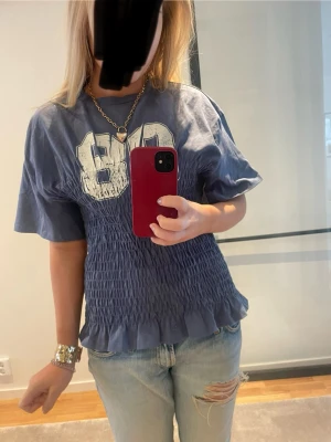 Blå smockad topp med tryck - Säljer en blå topp med stort vitt 80-tryck framtill. Toppen har smockad midja och volangkant nedtill, samt korta ärmar. Från Monki, storlek S, superbra skick. 