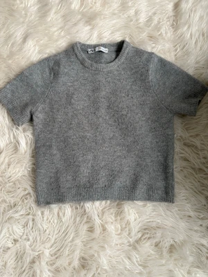 Grå stickad topp från Zara - Mysig grå stickad topp från Zara med korta ärmar och rund halsringning. Enkel och stilren design med ribbade kanter vid ärmslut och nederkant. Perfekt att matcha med jeans eller kjol för en chill look.