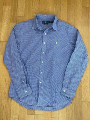 Blårutig skjorta från Ralph Lauren - Klassisk blårutig skjorta från Ralph Lauren med custom fit. Skjortan har lång ärm, knappar framtill och den ikoniska gula loggan broderad på bröstet. Tillverkad i mjuk bomull och perfekt för en clean och stilren look.